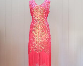 Peut inclure: Robe rose vif sans manches, longueur genou, avec un col en V et une bordure à franges. La robe est recouverte de paillettes et présente un motif orné et tourbillonnant sur le corsage. La robe est présentée sur un mannequin.