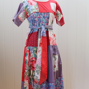 Puede incluir: Vestido midi patchwork con cuello redondo y mangas cortas. El vestido presenta una variedad de estampados florales y de lunares en rojo, azul, morado y blanco. Un cinturón de tela a juego ciñe la cintura, creando una silueta favorecedora.
