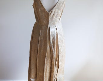 May include: A sleeveless floor-length dress in a shimmering champagne gold fabric with a tonal floral jacquard pattern. The garment features a deep V-neckline, a fitted bodice, and a full skirt with vertical pleats that extend to the hemline.