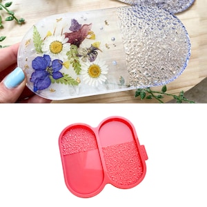 Può includere: Uno stampo in silicone a due pezzi per la colata di resina. Lo stampo è rosa e ha una superficie testurizzata. Ha la forma di una capsula con due scomparti separati.