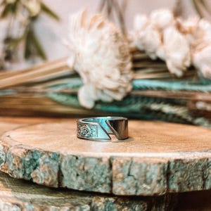 May include: A silver ring with a floral design on one side and a geometric cut on the other. The ring is displayed on a wooden surface with a natural edge, with dried flowers in the background. The ring is a unique piece of jewelry.