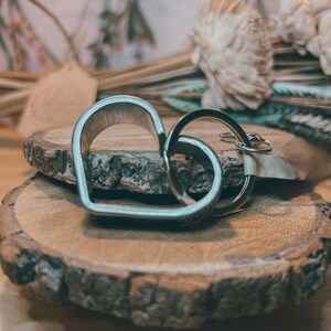 May include: A silver-colored heart-shaped keychain and a circular key ring are displayed on a wooden slice. The heart shape is a solid outline, and the key ring is attached to a small tag.