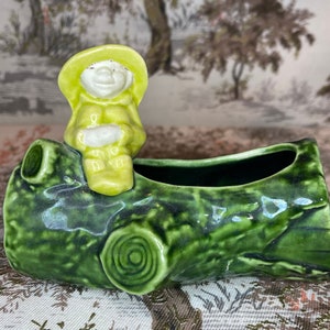 May include: A green ceramic planter shaped like a tree log with a small green figurine of a person sitting on top. The planter has a textured surface and a small hole in the top for drainage.
