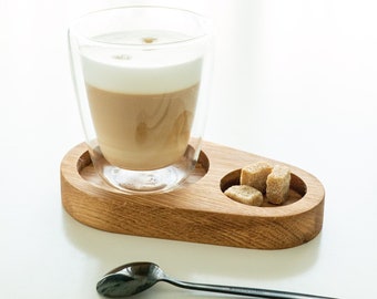 May include: A glass of latte with a layer of foam on top, served on a wooden tray with sugar cubes. A black metal spoon is on the white surface.