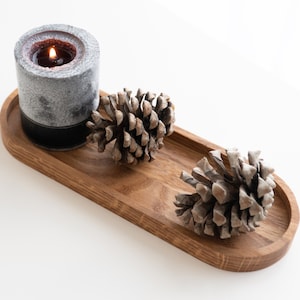 May include: A wooden tray with a gray candle and two pine cones. The candle is lit and the tray is oval shaped.