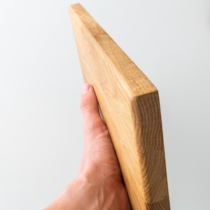 Puede incluir: Tabla de cortar rectangular de madera de color marr&oacute;n claro. La tabla se sostiene en &aacute;ngulo, mostrando su construcci&oacute;n gruesa y s&oacute;lida. La veta de la madera es visible, resaltando la textura natural del material. Mide unos 25 cm de largo.