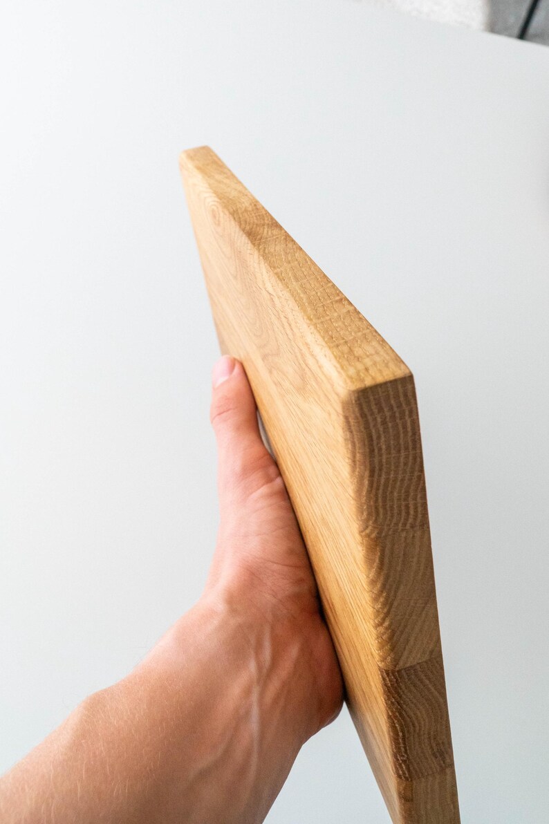 Puede incluir: Tabla de cortar rectangular de madera de color marr&oacute;n claro. La tabla se sostiene en &aacute;ngulo, mostrando su construcci&oacute;n gruesa y s&oacute;lida. La veta de la madera es visible, resaltando la textura natural del material. Mide unos 25 cm de largo.