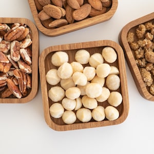 Puede incluir: Vista aérea de cuatro cuencos cuadrados de madera pequeños llenos de varios frutos secos. Los cuencos contienen almendras, nueces pecanas, nueces de macadamia y moras. Las nueces están dispuestas sobre una superficie blanca.