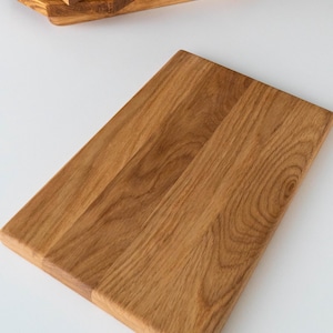 Puede incluir: Una tabla de cortar rectangular de madera con un acabado marr&oacute;n claro. La tabla es de madera y tiene una superficie lisa. Varias otras bandejas de madera est&aacute;n apiladas en el fondo. La tabla de cortar mide aproximadamente 30 cm de largo y 20 cm de ancho.