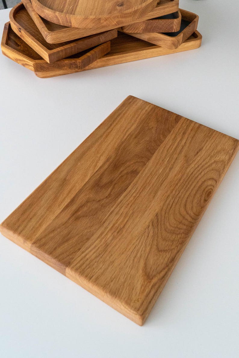 Puede incluir: Una tabla de cortar rectangular de madera con un acabado marr&oacute;n claro. La tabla es de madera y tiene una superficie lisa. Varias otras bandejas de madera est&aacute;n apiladas en el fondo. La tabla de cortar mide aproximadamente 30 cm de largo y 20 cm de ancho.