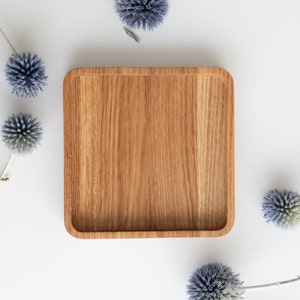 May include: A square wooden serving tray with rounded corners, showcasing a natural wood grain pattern. The tray is surrounded by blue thistle flowers on a white background. The tray is approximately 10 inches square.