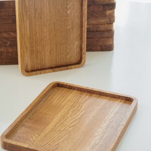 May include: A stack of square wooden serving trays, with one tray laying flat on a white surface. The trays are made of light brown wood with a natural grain pattern.