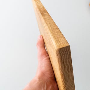 Puede incluir: Una tabla de cortar de madera clara con veta visible. La tabla es rectangular con un borde biselado. La tabla est&aacute; sujeta en la mano de una persona. La tabla mide aproximadamente 30 cm de largo.