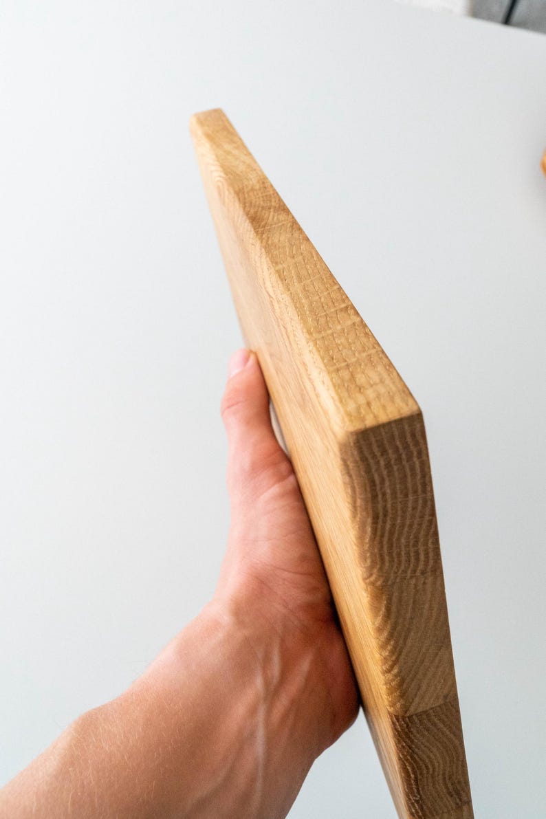 Puede incluir: Una tabla de cortar de madera clara con veta visible. La tabla es rectangular con un borde biselado. La tabla est&aacute; sujeta en la mano de una persona. La tabla mide aproximadamente 30 cm de largo.