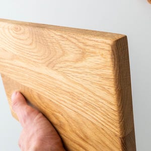 Puede incluir: Una tabla de cortar rectangular de madera clara con un borde biselado. La veta de la madera es visible y la tabla est&aacute; sujeta por una mano. La tabla probablemente se utiliza para la preparaci&oacute;n de alimentos.