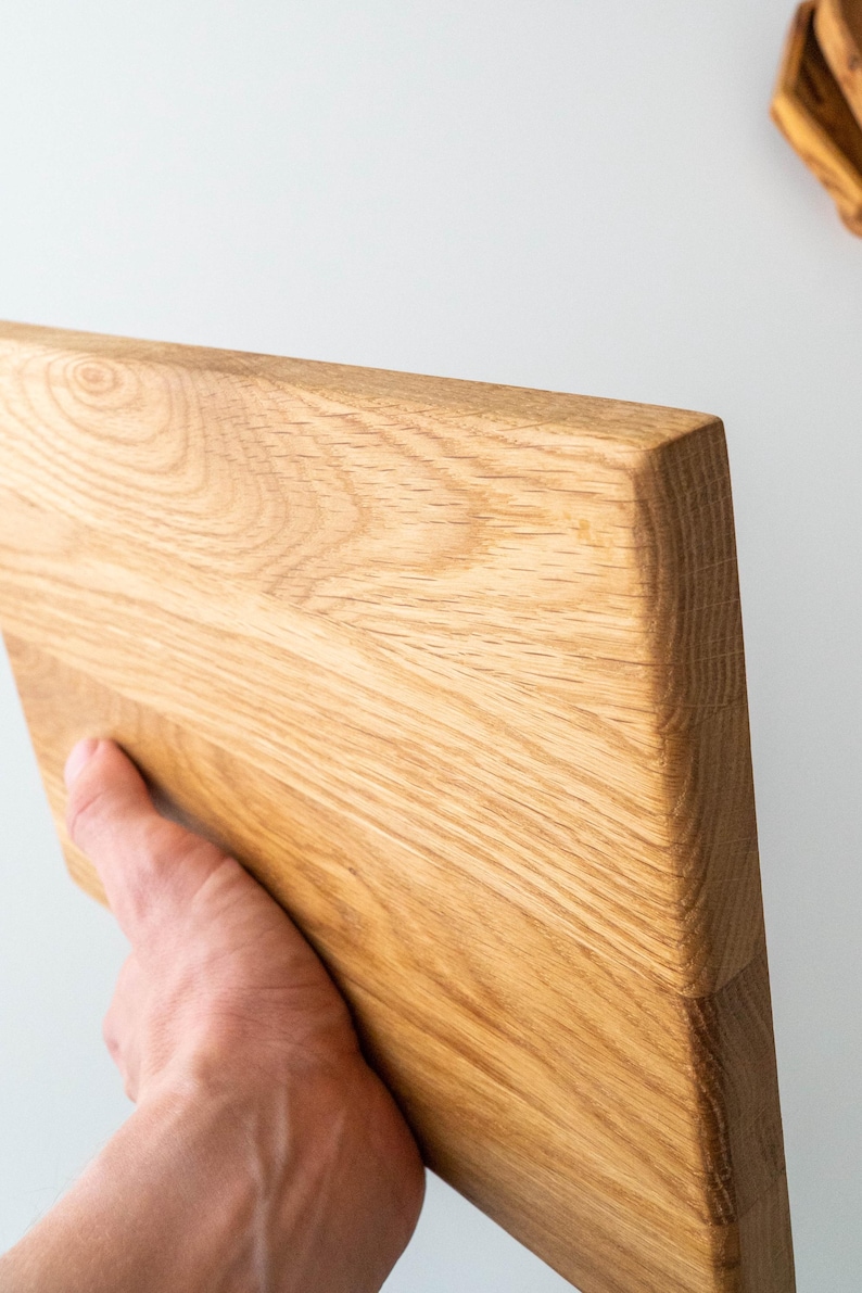 Puede incluir: Una tabla de cortar rectangular de madera clara con un borde biselado. La veta de la madera es visible y la tabla est&aacute; sujeta por una mano. La tabla probablemente se utiliza para la preparaci&oacute;n de alimentos.