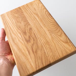 Puede incluir: Tabla de cortar rectangular de madera clara. Se aprecia la veta de la madera, y la tabla se sostiene en ángulo, mostrando su textura y color naturales. La tabla mide aproximadamente 30 cm de largo y 20 cm de ancho.