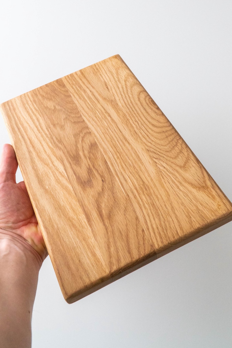 Puede incluir: Tabla de cortar rectangular de madera clara. Se aprecia la veta de la madera, y la tabla se sostiene en &aacute;ngulo, mostrando su textura y color naturales. La tabla mide aproximadamente 30 cm de largo y 20 cm de ancho.
