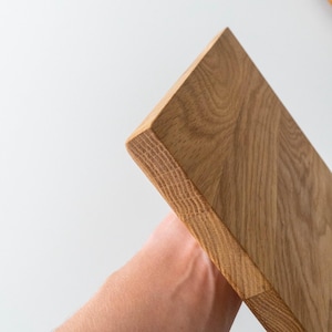 Puede incluir: Una tabla de cortar de madera marr&oacute;n claro con bordes redondeados. La veta de la madera es visible y la tabla se sostiene en &aacute;ngulo sobre un fondo blanco. La tabla parece nueva y sin usar.