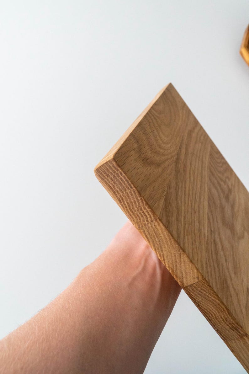 Puede incluir: Una tabla de cortar de madera marr&oacute;n claro con bordes redondeados. La veta de la madera es visible y la tabla se sostiene en &aacute;ngulo sobre un fondo blanco. La tabla parece nueva y sin usar.