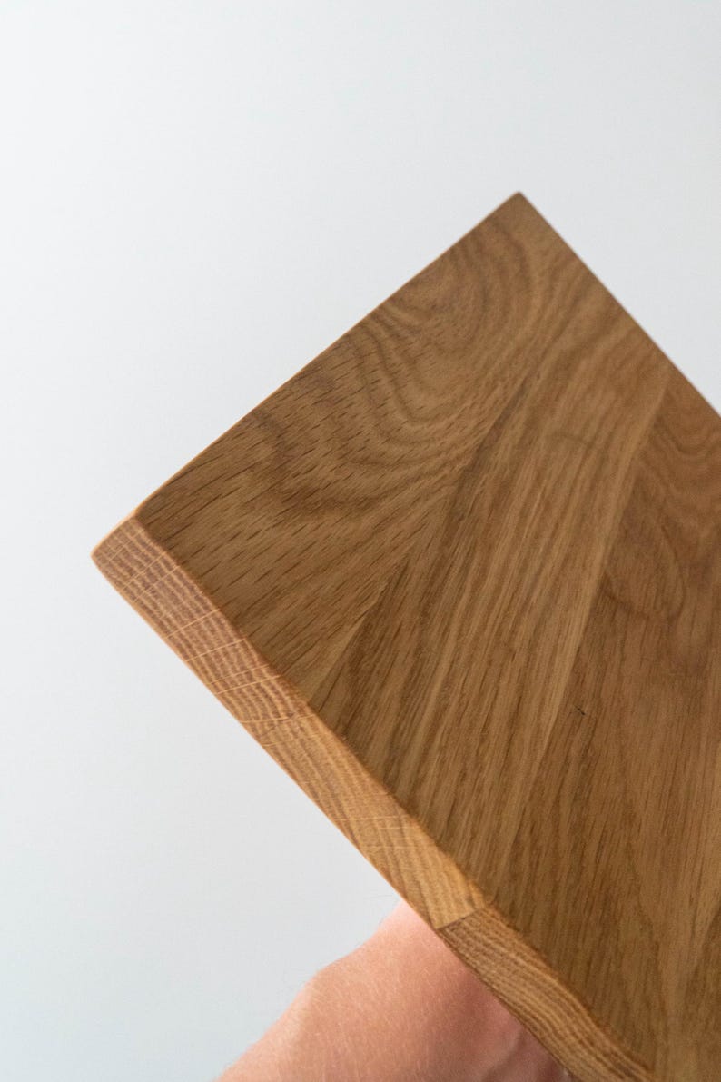 Puede incluir: Primer plano de una tabla de cortar de madera con veta natural. La tabla es cuadrada con un borde biselado. La madera es de color marr&oacute;n claro y tiene una textura suave. El fondo es blanco.
