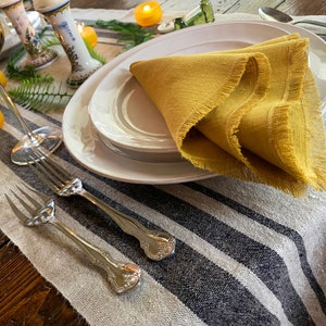 May include: A white dinner plate with a yellow linen napkin on top. The plate is set on a striped table runner with two silver forks.