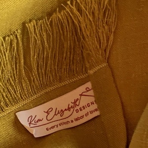 Peut inclure: Une couverture jaune moutarde avec une bordure en franges et une étiquette blanche qui indique "Kim Elizabeth Designs, Every stitch a labor of love."