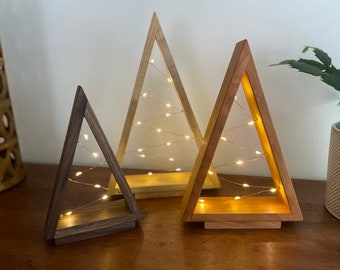 May include: Three wooden triangle-shaped Christmas tree decorations with warm white fairy lights inside. The trees are made of different wood tones, with the smallest tree being a dark brown wood and the two larger trees being a lighter brown wood.