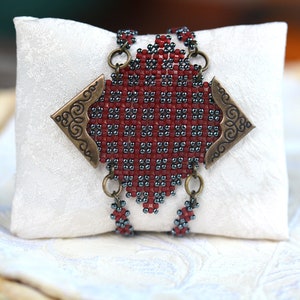 Puede incluir: Un brazalete de cuentas rojas y grises con un cierre de metal decorativo. El brazalete está sobre una almohada blanca.