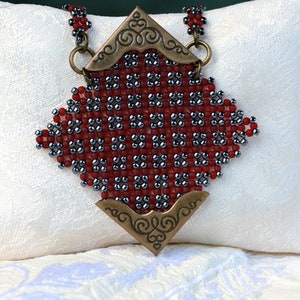 Może przedstawiać: Naszyjnik z czerwonych i srebrnych koralików z ozdobnym metalowym wisiorem. Wisiorek ma kształt diamentu i zdobi go kwiatowy wzór.