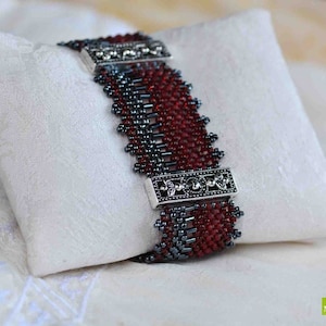 Könnte beinhalten: Ein rotes und graues Perlenarmband mit silberfarbenen Dekor-Akzenten. Das Armband liegt auf einer weißen Stoffoberfläche.