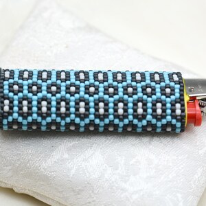 Könnte beinhalten: Ein schwarzes, weißes und blaues Perlen-Feuerzeugetui mit geometrischem Muster. Das Etui besteht aus kleinen Perlen und hat einen Reißverschluss.