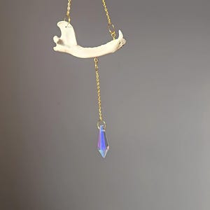 May include: A unique, handcrafted pendant featuring a bone jaw suspended from a delicate gold chain. A faceted, iridescent crystal hangs below the jaw. The pendant is displayed against a gray background.