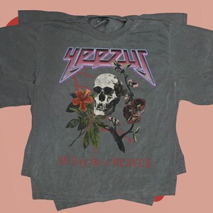 May include: A gray t-shirt with a skull and flowers graphic. The text "YEEZY" is printed in a large, bold font above the graphic. The text "All Love is Heavy" is printed below the graphic.
