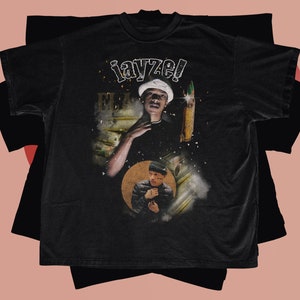 May include: Black t-shirt featuring a graphic print of a man wearing a hat and holding a microphone. The text "iayze!" is printed on the shirt.