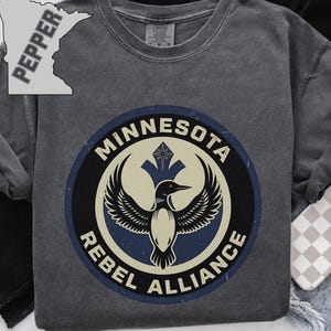 Könnte beinhalten: Ein anthrazitfarbenes Kurzarm-T-Shirt mit einem kreisförmigen Motiv, das einen Eistaucher, den Text Minnesota Rebel Alliance und einen Stern zeigt. Eine kleine graue Karte von Minnesota mit dem Wort Pepper befindet sich oben links.