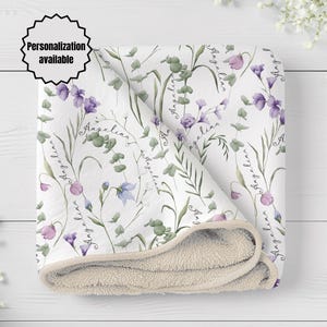 Könnte beinhalten: Eine weiße Decke mit einem sich wiederholenden Muster aus grünen Eukalyptusblättern und violetten Blumen, wobei der Name Angelina in Schreibschrift über das Design verteilt ist. Die Decke hat eine weiche, strukturierte beige Unterseite.