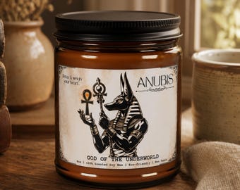 May include: A brown glass candle jar with a black lid and a white label featuring a black illustration of the Egyptian god Anubis holding an ankh. The label text reads Anubis, God of the Underworld, and Relax & weigh your heart, along with soy wax details.