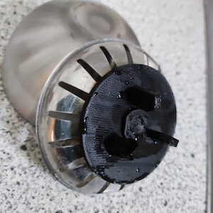 Puede incluir: Primer plano de un colador de fregadero plateado con un tapón de plástico negro. El colador tiene un diseño circular con múltiples agujeros y un borde elevado. El tapón es redondo y tiene una pequeña manija.