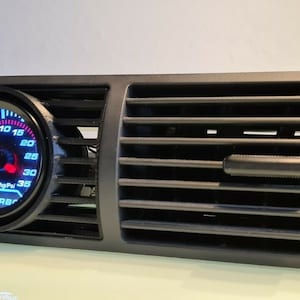 Puede incluir: Ventilación de aire de coche de plástico negro con un manómetro redondo que indica "TURBO" y una aguja roja que apunta a 15. El manómetro tiene una esfera azul con números blancos y un bisel negro.