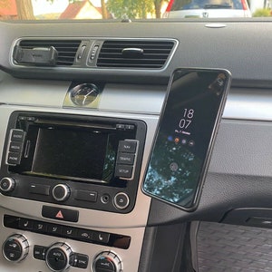 Könnte beinhalten: Ein schwarzes Smartphone mit einem dunklen Bildschirm, auf dem die Uhrzeit 18:07 angezeigt wird, ist im Armaturenbrett eines Autos montiert. Das Armaturenbrett des Autos ist grau und hat Radio- und Klimasteuerknöpfe.