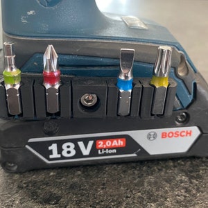 May include: A black and blue Bosch drill with a 18V 2.0Ah Li-Ion battery. The drill has a bit holder with four different bits, including a Phillips head, a flat head, a star head, and a square head.