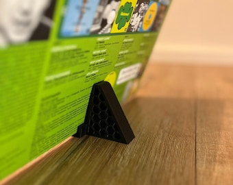 May include: A black triangular record stand with a honeycomb pattern on the front. The stand is holding up a vinyl record with a green and yellow cover.