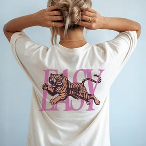 May include: Cream-colored t-shirt with a pink graphic of a tiger and the word "EASY". The tiger is brown and orange with black stripes. The shirt has short sleeves and a crew neck.