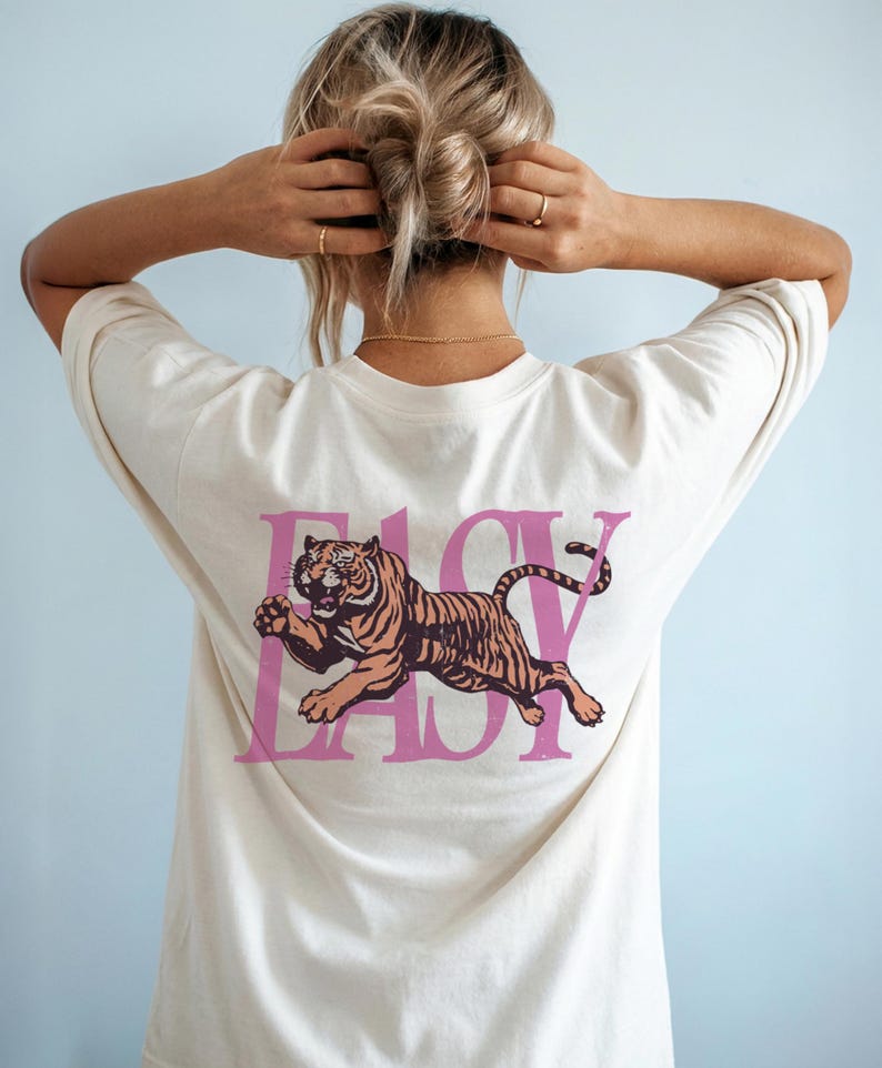 May include: Cream-colored t-shirt with a pink graphic of a tiger and the word "EASY". The tiger is brown and orange with black stripes. The shirt has short sleeves and a crew neck.