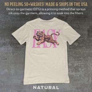 May include: Cream-colored t-shirt with a pink and orange tiger graphic and the word "EASY" in pink. The shirt has the text "NO PEELING 50+WASHES! MADE & SHIPS IN THE USA" at the top and "NATURAL" at the bottom.