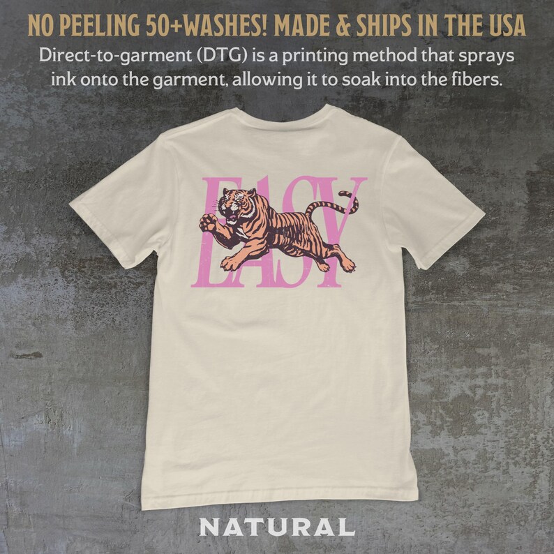 May include: Cream-colored t-shirt with a pink and orange tiger graphic and the word "EASY" in pink. The shirt has the text "NO PEELING 50+WASHES! MADE & SHIPS IN THE USA" at the top and "NATURAL" at the bottom.