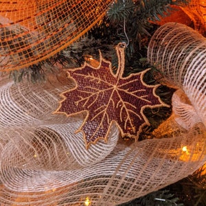 May include: A close-up of a fall-themed decoration. A burgundy leaf-shaped ornament with gold trim hangs from a silver hook. The ornament is surrounded by orange and beige mesh ribbon, and green pine branches. Warm lights add a festive glow.