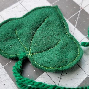May include: A green felt leaf with a green yarn tail. The leaf has a light green stitch around the edge and a light green stitch outlining the veins of the leaf.