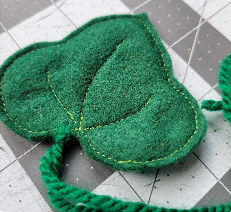 May include: A green felt leaf with a green yarn tail. The leaf has a light green stitch around the edge and a light green stitch outlining the veins of the leaf.